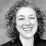 devon hase, black and white picture, curly shoulder length hair, black blazer, smiling.