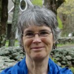 Rebecca Bradshaw, with grey hair and glasses, blue jacket, standing in front of a stone wall and trees and building.
