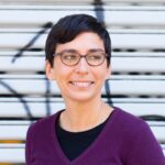 Tara Mulay, short dark hair and glasses ,looking to the right, purple v-neck sweater over a black shirt, standing in front of a series of white slats.