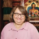 Sharon Salzberg, has glasses and wearing a pink blouse, standing in front of a wall with a Buddhist image.