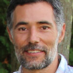 Alexis Santos, a man with short dark gray hair, a beard, and wearing a checkered shirt, standing against a tree trunk.