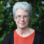 Andrea Fella,a woman with short gray hair and glasses, dressed in an orange top and black cardigan, with a backdrop of dark green foliage. 